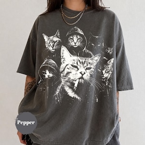 K&ouml;nnte beinhalten: Ein anthrazitfarbenes Kurzarm-T-Shirt mit einem gro&szlig;en wei&szlig;en Grafikdruck von mehreren Katzen, darunter eine mit Kapuzenpullover und eine weitere mit einer Zigarette im Mund. Das Wort Pepper ist in Wei&szlig; auf einem grauen kreisf&ouml;rmigen Hintergrund gedruckt.