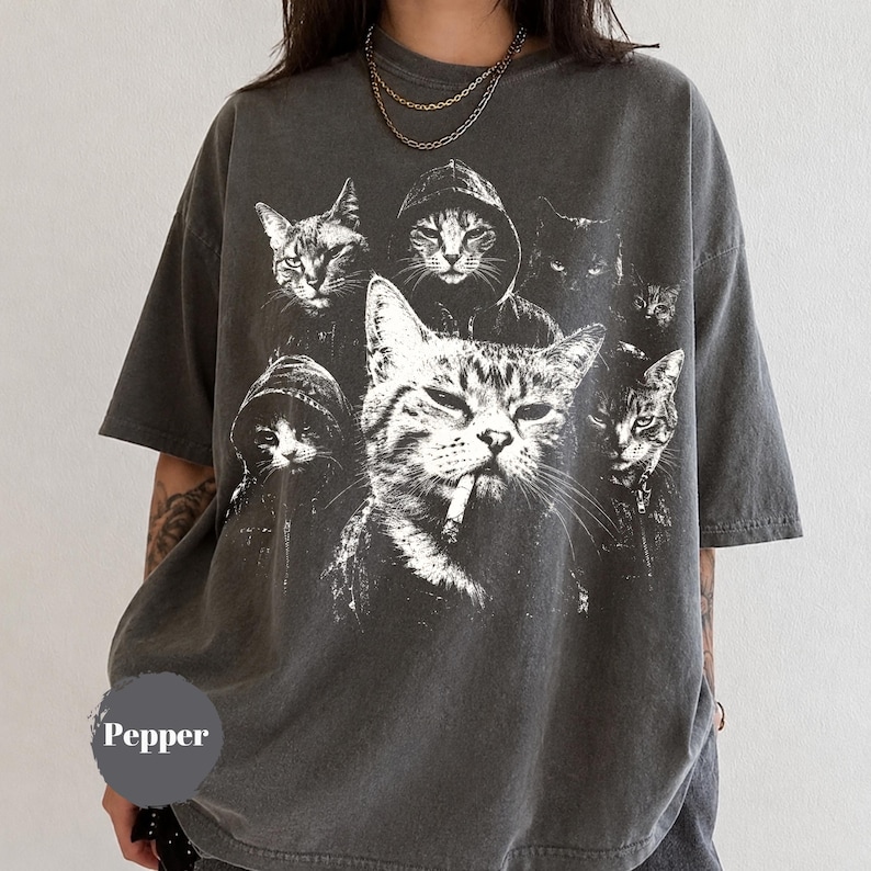 K&ouml;nnte beinhalten: Ein anthrazitfarbenes Kurzarm-T-Shirt mit einem gro&szlig;en wei&szlig;en Grafikdruck von mehreren Katzen, darunter eine mit Kapuzenpullover und eine weitere mit einer Zigarette im Mund. Das Wort Pepper ist in Wei&szlig; auf einem grauen kreisf&ouml;rmigen Hintergrund gedruckt.