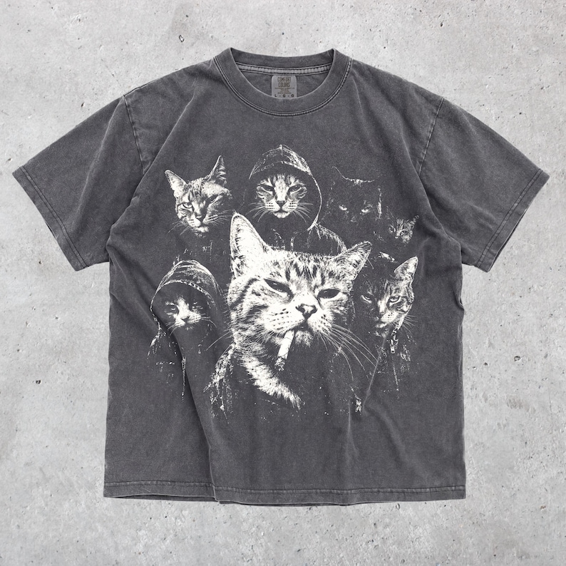 K&ouml;nnte beinhalten: Ein anthrazitfarbenes Kurzarm-T-Shirt mit einem wei&szlig;en Grafikdruck von mehreren Katzen. Die zentrale Katze ist mit einer Zigarette im Mund dargestellt, w&auml;hrend andere Katzen Kapuzenpullover tragen, was ein geschichtetes, monochromes Design ergibt.