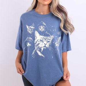K&ouml;nnte beinhalten: Ein staubblaues Kurzarm-T-Shirt mit einem gro&szlig;en, verwaschenen wei&szlig;en Grafikdruck von mehreren Katzen. Die zentrale Katze hat eine Zigarette im Mund und ist von anderen Katzen umgeben, von denen einige Kapuzenpullover tragen, in einem monochromen Stil.