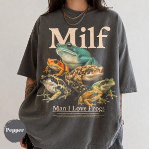 Puede incluir: Una camiseta de manga corta gris carbón con un estampado gráfico de siete ranas variadas. El texto "Milf" aparece en la parte superior en una fuente grande de color crema, con "Man I Love Frogs" y un pequeño párrafo impreso debajo.