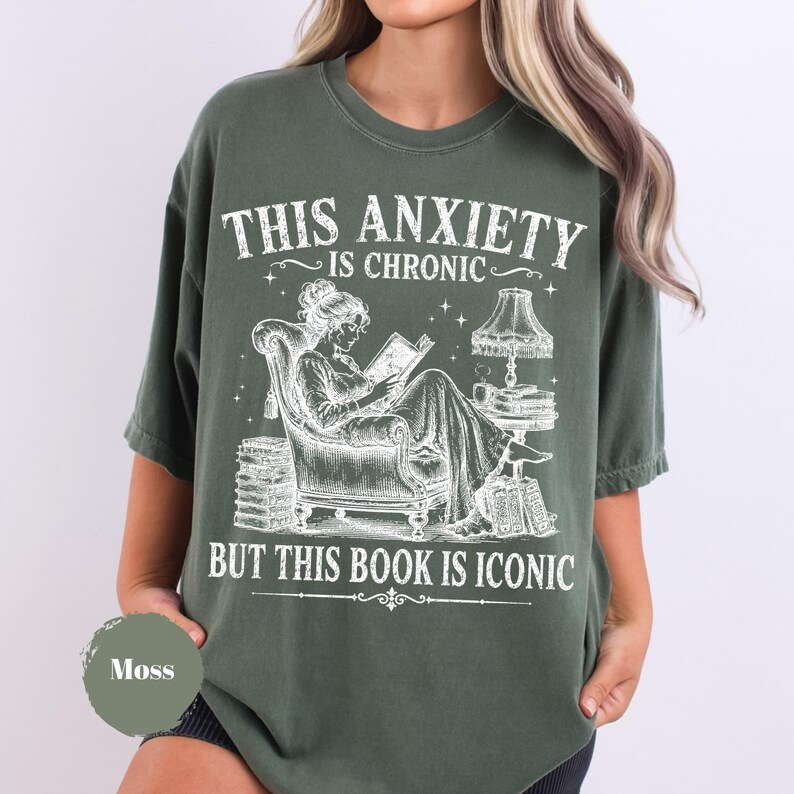 May include: A moss green short-sleeve t-shirt featuring a white graphic illustration of a person reading in an armchair with stacks of books and a lamp. The text reads: THIS ANXIETY IS CHRONIC BUT THIS BOOK IS ICONIC. The shirt has a crew neckline.