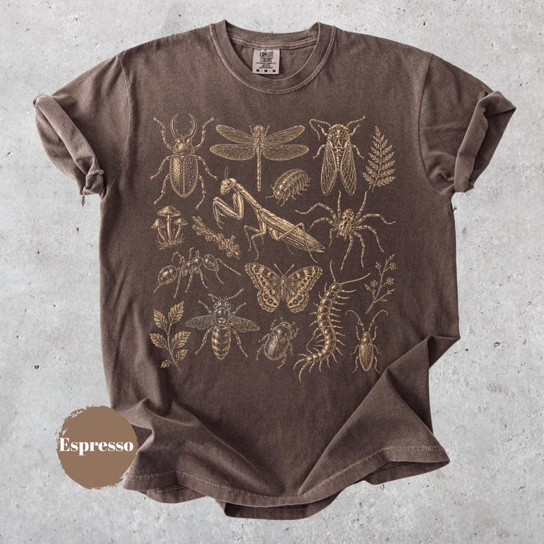 May include: A dark brown short-sleeve t-shirt featuring a detailed gold-toned graphic print of various insects, including a beetle, dragonfly, mantis, butterfly, and centipede, alongside small botanical elements like mushrooms and fern leaves.
