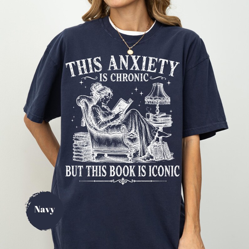 May include: A navy blue t-shirt featuring a white graphic of a person reading in an armchair next to a lamp and stacks of books. The text reads: THIS ANXIETY IS CHRONIC BUT THIS BOOK IS ICONIC. The design includes stars and decorative lines.