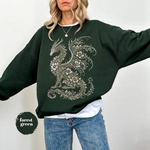 May include: A forest green crewneck sweatshirt featuring a large, intricate graphic of a dragon composed entirely of detailed floral patterns and vines in shades of cream and light gray, worn over a white undershirt and blue denim jeans.