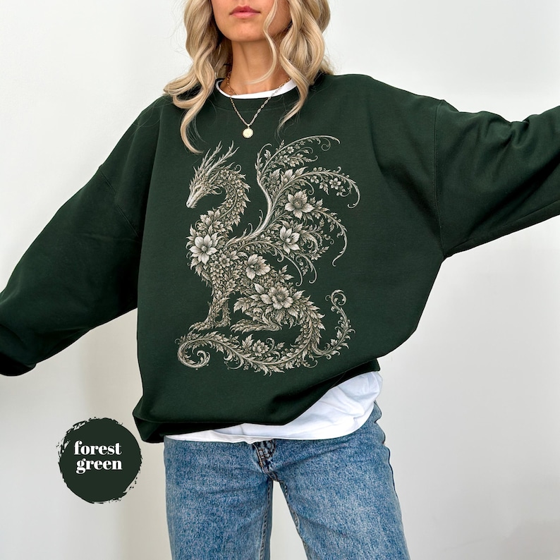 May include: A forest green crewneck sweatshirt featuring a large, intricate graphic of a dragon composed entirely of detailed floral patterns and vines in shades of cream and light gray, worn over a white undershirt and blue denim jeans.