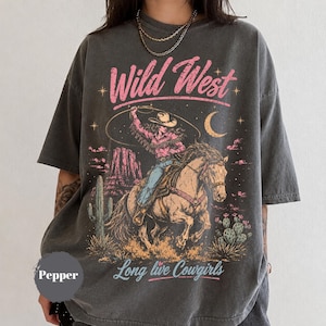 May include: A charcoal grey short-sleeve t-shirt featuring a graphic print of a cowgirl riding a horse with a lasso. The design includes pink text reading Wild West and Long live Cowgirls, a crescent moon, stars, cacti, and desert rock formations.