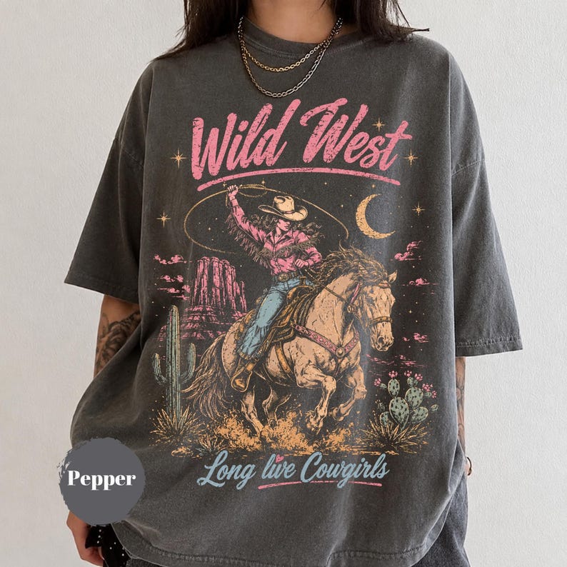 May include: A charcoal grey short-sleeve t-shirt featuring a graphic print of a cowgirl riding a horse with a lasso. The design includes pink text reading Wild West and Long live Cowgirls, a crescent moon, stars, cacti, and desert rock formations.