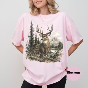 K&ouml;nnte beinhalten: Ein hellrosa T-Shirt mit kurzen &Auml;rmeln und einem detaillierten Grafikdruck eines Hirsches in einer Waldlandschaft mit Bergen und einem See im Hintergrund. Das Wort Blossom ist in Schwarz in der unteren rechten Ecke des T-Shirts aufgedruckt.