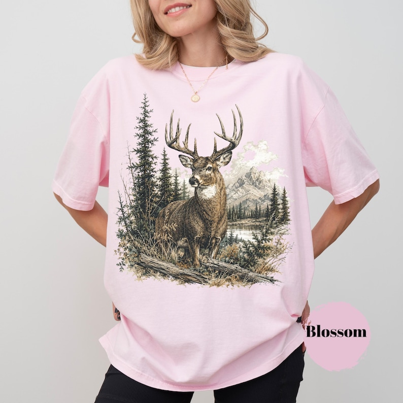 K&ouml;nnte beinhalten: Ein hellrosa T-Shirt mit kurzen &Auml;rmeln und einem detaillierten Grafikdruck eines Hirsches in einer Waldlandschaft mit Bergen und einem See im Hintergrund. Das Wort Blossom ist in Schwarz in der unteren rechten Ecke des T-Shirts aufgedruckt.