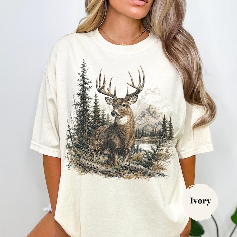 K&ouml;nnte beinhalten: Ein elfenbeinfarbenes Kurzarm-T-Shirt mit einem detaillierten Grafikdruck eines Hirsches, der in einem Wald mit Kiefern und Bergen im Hintergrund steht. Die Illustration ist in erdigen Braun-, Gr&uuml;n- und Graut&ouml;nen gehalten und mittig platziert.