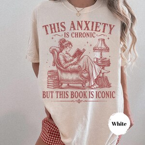 May include: A cream-colored t-shirt featuring a red graphic illustration of a person reading a book in an armchair. The text reads, THIS ANXIETY IS CHRONIC BUT THIS BOOK IS ICONIC. The design includes a lamp, stacks of books, and small stars.