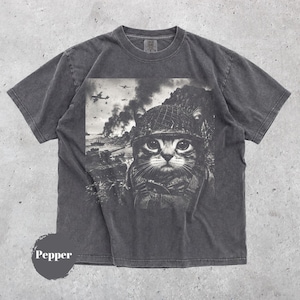 Peut inclure: Un t-shirt gris anthracite à manches courtes avec une impression graphique en noir et blanc représentant un chat portant un casque militaire. L'arrière-plan montre une scène de champ de bataille avec de la fumée, des avions et des chars. Le mot Pepper est imprimé en bas.