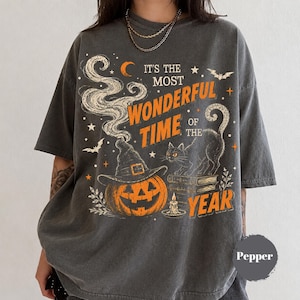 May include: A charcoal grey t-shirt featuring a graphic print with the text IT'S THE MOST WONDERFUL TIME OF THE YEAR. The design includes a jack-o'-lantern wearing a witch hat, a black cat, a stack of books, a candle, bats, stars, and a crescent moon.