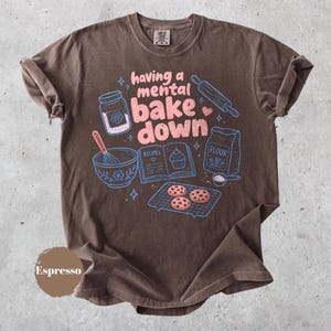 May include: A dark brown short-sleeve t-shirt featuring a graphic print with the text "having a mental bake down" in pink. The design includes blue line art of a jar, rolling pin, mixing bowl with whisk, recipe book, flour bag, and cookies.