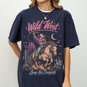 May include: A navy blue t-shirt featuring a graphic print of a cowgirl riding a horse in a desert landscape. The design includes the text "Wild West" in pink and "Long live Cowgirls" in light blue, with cacti, mesas, and a crescent moon.