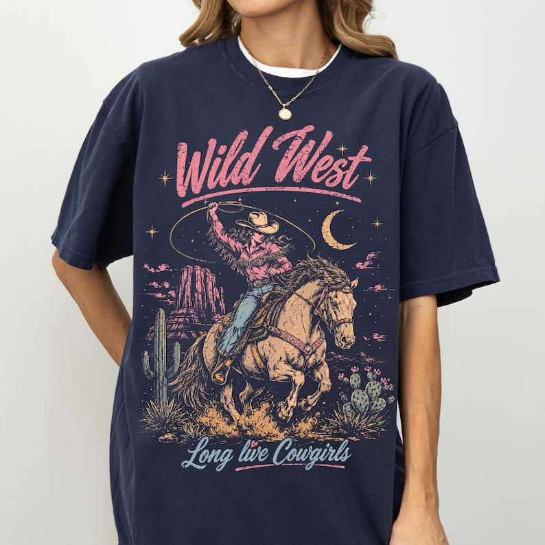 May include: A navy blue t-shirt featuring a graphic print of a cowgirl riding a horse in a desert landscape. The design includes the text "Wild West" in pink and "Long live Cowgirls" in light blue, with cacti, mesas, and a crescent moon.