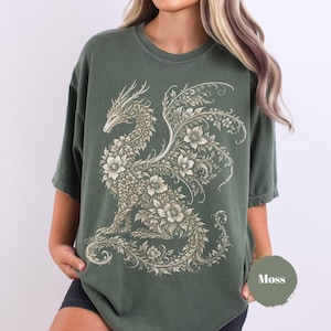 May include: A moss green short-sleeve t-shirt featuring a large, intricate graphic of a dragon composed entirely of floral and leafy patterns in a light cream color. The dragon is depicted in profile with detailed wings and a long, curled tail.