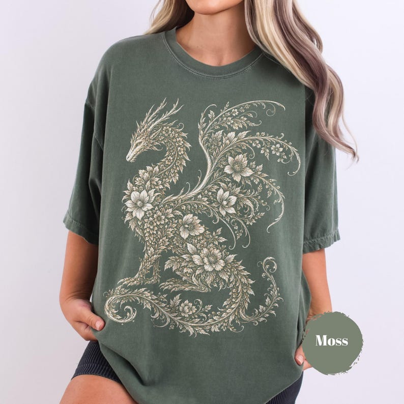 May include: A moss green short-sleeve t-shirt featuring a large, intricate graphic of a dragon composed entirely of floral and leafy patterns in a light cream color. The dragon is depicted in profile with detailed wings and a long, curled tail.