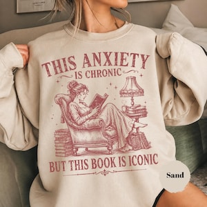 May include: A sand-colored crewneck sweatshirt featuring a maroon graphic of a person sitting in an armchair reading a book. The text reads: THIS ANXIETY IS CHRONIC BUT THIS BOOK IS ICONIC. The illustration includes a lamp, stacks of books, and stars.