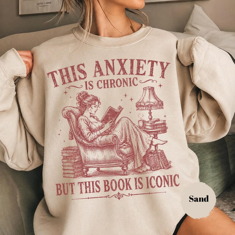 May include: A sand-colored crewneck sweatshirt featuring a maroon graphic of a person sitting in an armchair reading a book. The text reads: THIS ANXIETY IS CHRONIC BUT THIS BOOK IS ICONIC. The illustration includes a lamp, stacks of books, and stars.