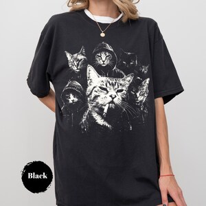 K&ouml;nnte beinhalten: Ein schwarzes kurz&auml;rmeliges T-Shirt mit einem wei&szlig;en Grafikdruck von mehreren Katzen, darunter eine zentrale Katze mit einer Zigarette im Mund und andere, die Kapuzenpullover tragen. Das T-Shirt hat einen Rundhalsausschnitt und eine lockere Passform.