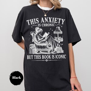 May include: A black short-sleeve t-shirt featuring a white graphic of a person reading in an armchair. The text reads THIS ANXIETY IS CHRONIC BUT THIS BOOK IS ICONIC. The design includes a lamp, stacks of books, and stars in a detailed line art style.
