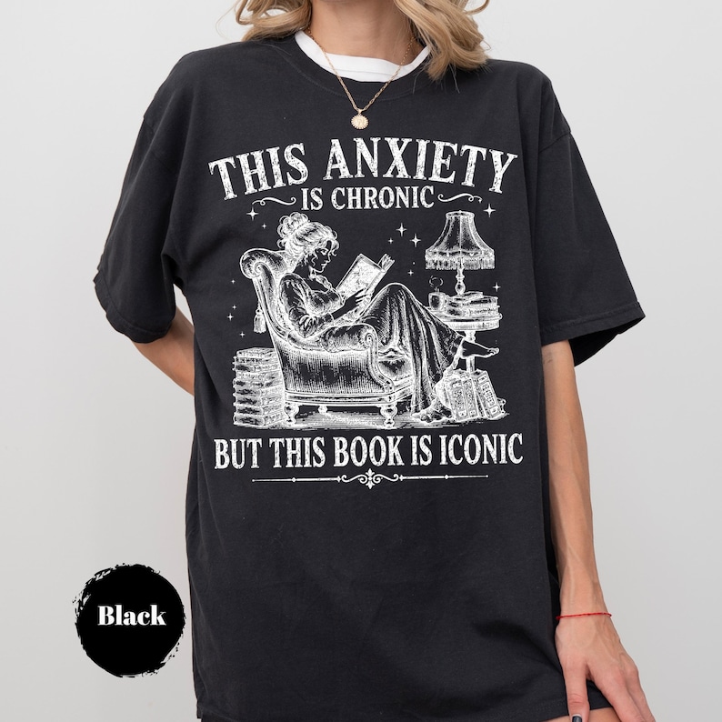 May include: A black short-sleeve t-shirt featuring a white graphic of a person reading in an armchair. The text reads THIS ANXIETY IS CHRONIC BUT THIS BOOK IS ICONIC. The design includes a lamp, stacks of books, and stars in a detailed line art style.