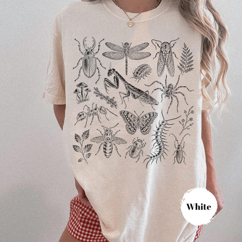 May include: A cream-colored t-shirt featuring a detailed black ink illustration of various insects, including a praying mantis, butterfly, beetle, and spider, alongside small botanical elements like mushrooms and fern leaves.