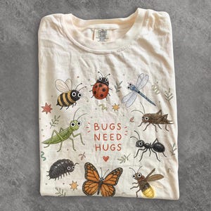 May include: A cream-coloured t-shirt featuring a graphic print of various insects including a bee, ladybird, dragonfly, grasshopper, ant, woodlouse, butterfly, and firefly. The text BUGS NEED HUGS is printed in the centre in a reddish-brown font.