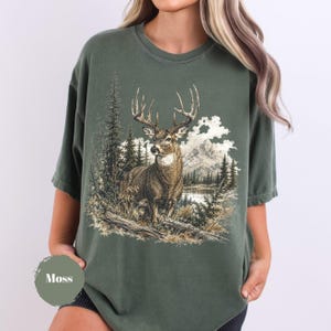 K&ouml;nnte beinhalten: Ein moosgr&uuml;nes T-Shirt mit Rundhalsausschnitt und einem detaillierten Grafikdruck eines Hirsches mit gro&szlig;em Geweih, der in einer Waldlandschaft mit Kiefern, hohem Gras und Bergen im Hintergrund steht. Das Design ist in erdigen Braun- und Wei&szlig;t&ouml;nen gehalten.