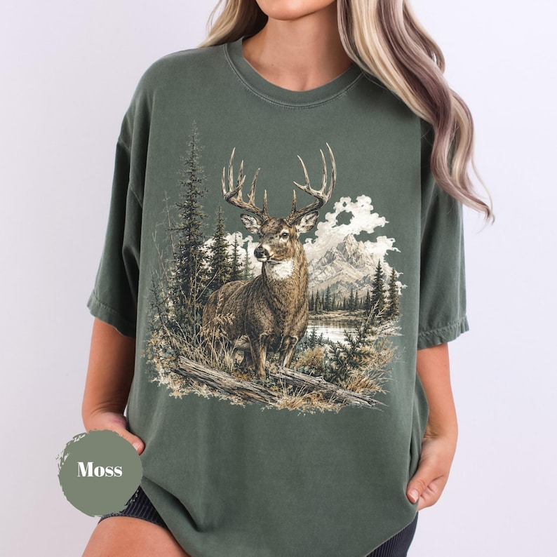 K&ouml;nnte beinhalten: Ein moosgr&uuml;nes T-Shirt mit Rundhalsausschnitt und einem detaillierten Grafikdruck eines Hirsches mit gro&szlig;em Geweih, der in einer Waldlandschaft mit Kiefern, hohem Gras und Bergen im Hintergrund steht. Das Design ist in erdigen Braun- und Wei&szlig;t&ouml;nen gehalten.