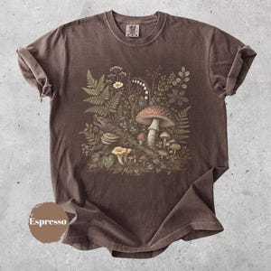 May include: A brown short-sleeve t-shirt featuring a detailed botanical illustration of various mushrooms, ferns, and wildflowers. The graphic is centered on the chest and includes a variety of fungi species. A circular tag reads Espresso.