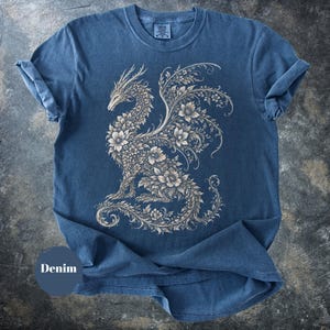 May include: A blue short-sleeved t-shirt featuring a large, intricate graphic of a dragon composed entirely of floral and vine patterns in a light beige color. The shirt has a crew neckline and a visible brand tag on the inside of the collar.