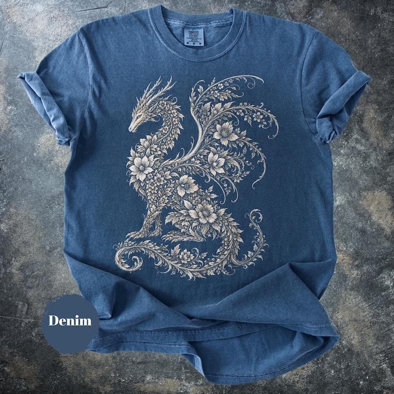 May include: A blue short-sleeved t-shirt featuring a large, intricate graphic of a dragon composed entirely of floral and vine patterns in a light beige color. The shirt has a crew neckline and a visible brand tag on the inside of the collar.