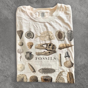 May include: A cream-coloured t-shirt featuring a detailed graphic print of various fossils. The design includes illustrations of a dinosaur skull, fish skeleton, and shells with the text FOSSILS HISTORY IN THE ROCKS printed in the centre.