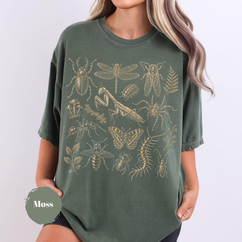 May include: A moss green short-sleeve t-shirt featuring a gold-toned graphic print of various insects, including a beetle, dragonfly, praying mantis, spider, butterfly, and centipede, alongside botanical elements like mushrooms and ferns.