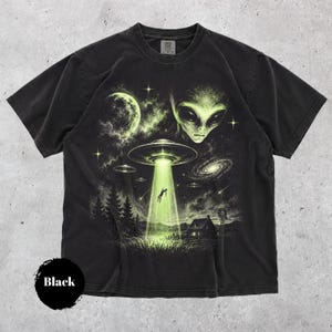 Może przedstawiać: Czarny t-shirt z krótkim rękawem z nadrukiem przedstawiającym dużą głowę kosmity, księżyc i kilka latających spodków na tle rozgwieżdżonego nieba. Zielona wiązka światła unosi postać nad domem, drzewami i wiatrakiem w polu.
