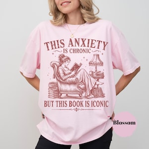 May include: A light pink t-shirt featuring a dark red graphic of a person reading in an armchair. The text reads: THIS ANXIETY IS CHRONIC BUT THIS BOOK IS ICONIC. The illustration includes a lamp, stacks of books, and stars surrounding the central figure.