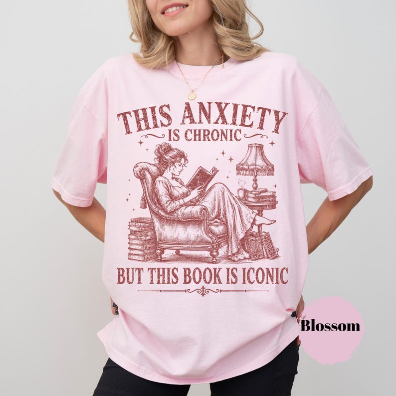 May include: A light pink t-shirt featuring a dark red graphic of a person reading in an armchair. The text reads: THIS ANXIETY IS CHRONIC BUT THIS BOOK IS ICONIC. The illustration includes a lamp, stacks of books, and stars surrounding the central figure.