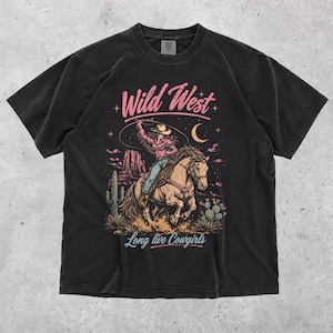 May include: A black short-sleeved t-shirt featuring a graphic print of a cowgirl riding a horse with a lasso. The text reads Wild West at the top and Long live Cowgirls at the bottom. The design includes a desert landscape with cacti and a moon.