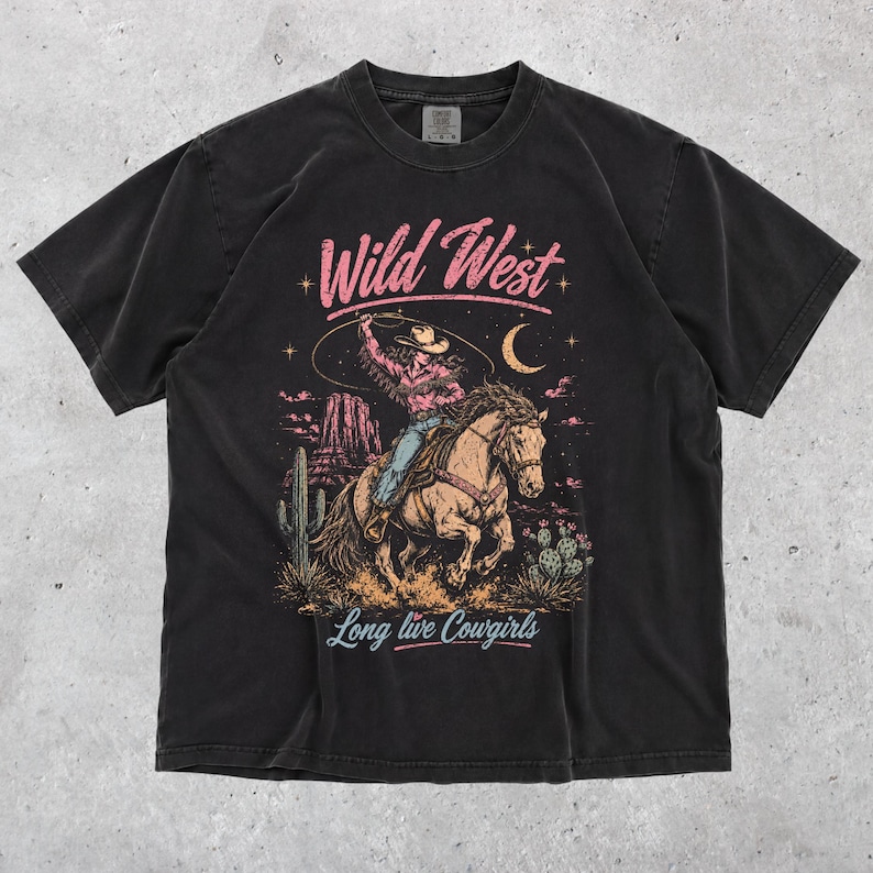 May include: A black short-sleeved t-shirt featuring a graphic print of a cowgirl riding a horse with a lasso. The text reads Wild West at the top and Long live Cowgirls at the bottom. The design includes a desert landscape with cacti and a moon.