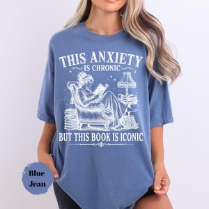May include: A blue jean colored t-shirt featuring a white graphic of a person reading in an armchair surrounded by stacks of books and a lamp. The text reads: THIS ANXIETY IS CHRONIC BUT THIS BOOK IS ICONIC. The design includes stars and decorative lines.