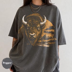 May include: A charcoal gray short-sleeve t-shirt featuring a large graphic print of a bison head centered on the chest. The artwork includes a golden sun, a mountain range, and a forest landscape. The word Pepper is printed in a small gray circle.