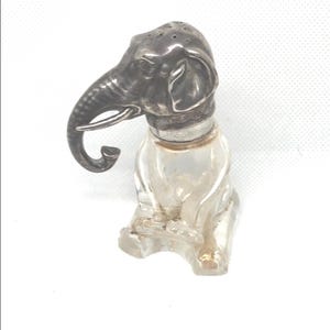 May include: A vintage salt shaker in the shape of an elephant. The elephant's head is silver-colored, with a detailed trunk and small holes on top. The body is clear glass, and the shaker is sitting upright.
