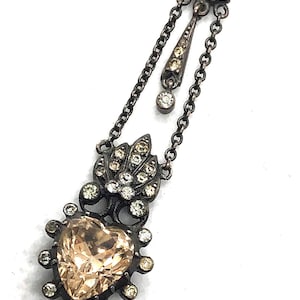 May include: Antique silver-tone necklace featuring a heart-shaped, peach-colored gemstone pendant. The pendant is surrounded by small, clear gemstones. The necklace has a delicate chain and additional decorative elements with clear and yellow gemstones.