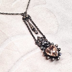 May include: A dark metal necklace featuring a central heart-shaped peach-coloured gemstone surrounded by small clear stones. The pendant is suspended from a delicate chain and includes an ornate decorative element with additional embedded crystals.