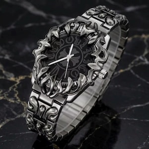 May include: A silver-toned wristwatch with an ornate, dark gray face and matching band. The watch face features a decorative pattern and silver hands. The band and bezel have a detailed, sculpted design. The watch is displayed on a dark, marbled surface.