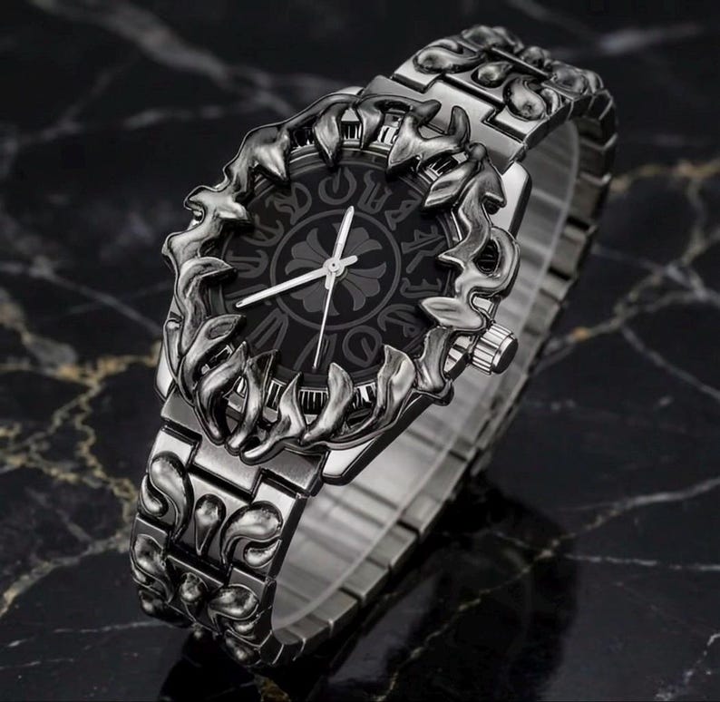 May include: A silver-toned wristwatch with an ornate, dark gray face and matching band. The watch face features a decorative pattern and silver hands. The band and bezel have a detailed, sculpted design. The watch is displayed on a dark, marbled surface.