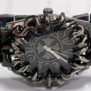 May include: Close-up of a dark gray wristwatch with a decorative, ornate bezel. The watch face is gray with a floral design and silver hands. The watch band is a dark gray, linked design. The watch has a silver crown.